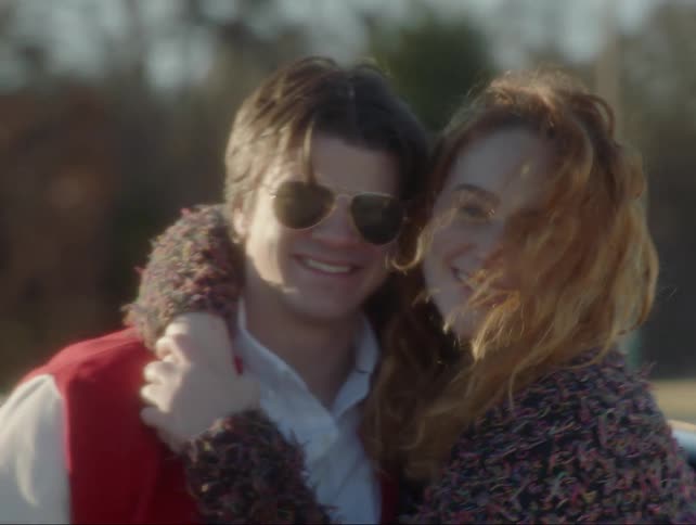 A teenage couple are hugging and smiling as they stand outside during the day