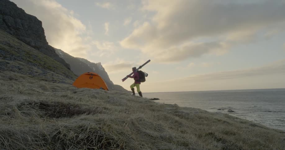 Woman carrying skis up a hill toward tent overlooking the ocean at dusk