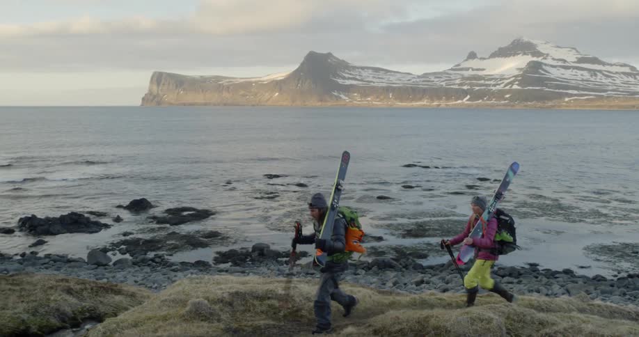 Two people carrying skis hike along the shore at sunset