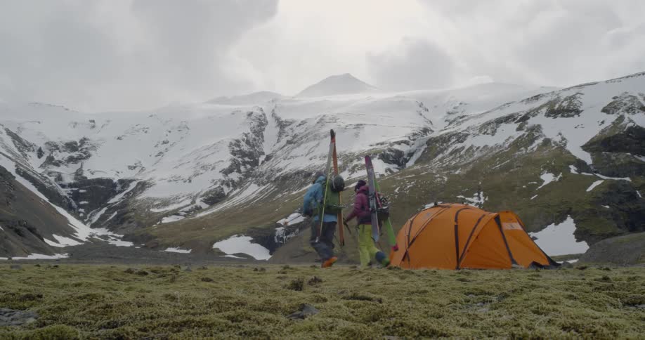 A man and woman have skis strapped to their backs in snowy mountains