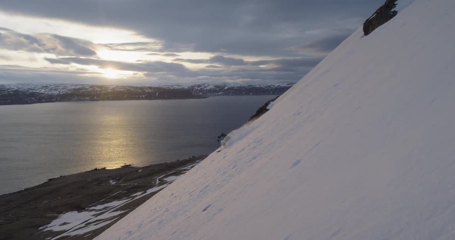 A skier glides down a steep mountainside at sunset with ocean behind
