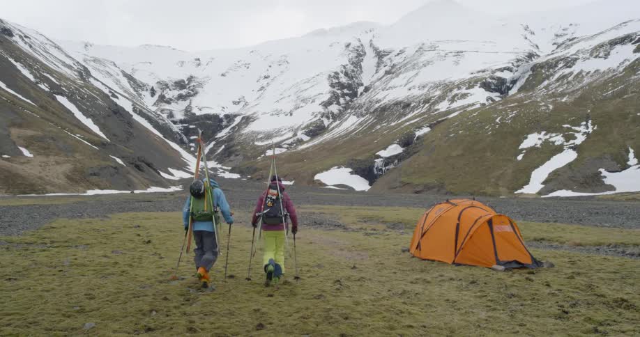 Two people hike towards a tent with skis on their back