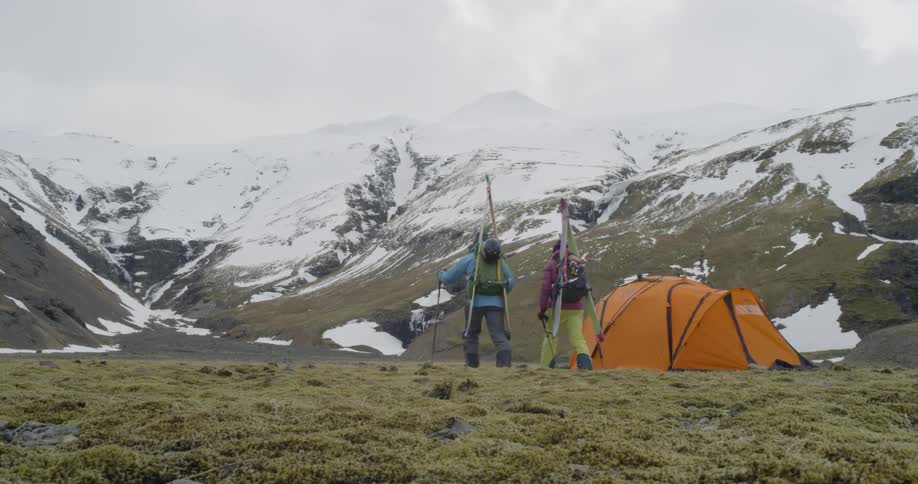 A couple carrying their skis and packs walk past a tent up a mountain