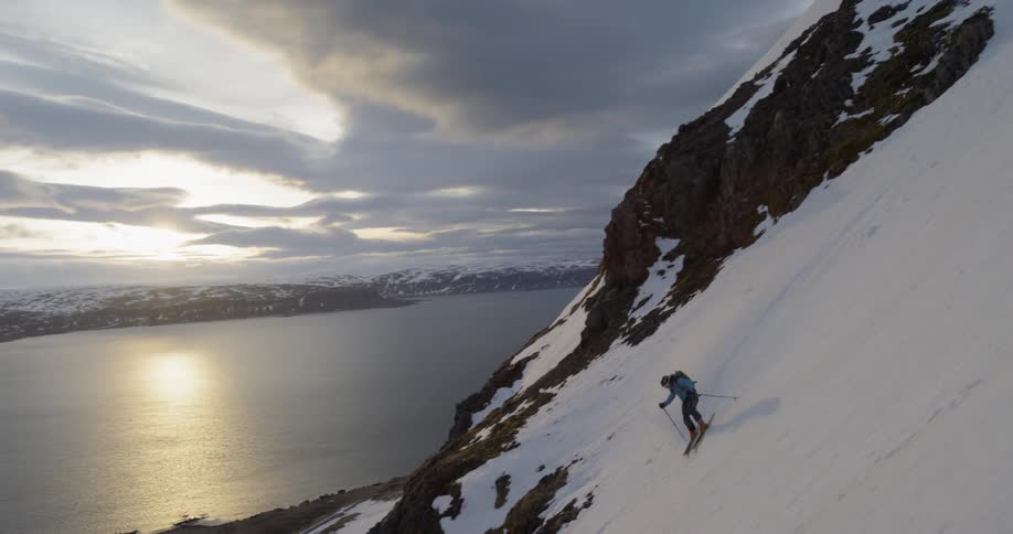 A skier turns down a snowy mountain side towards water at sunset