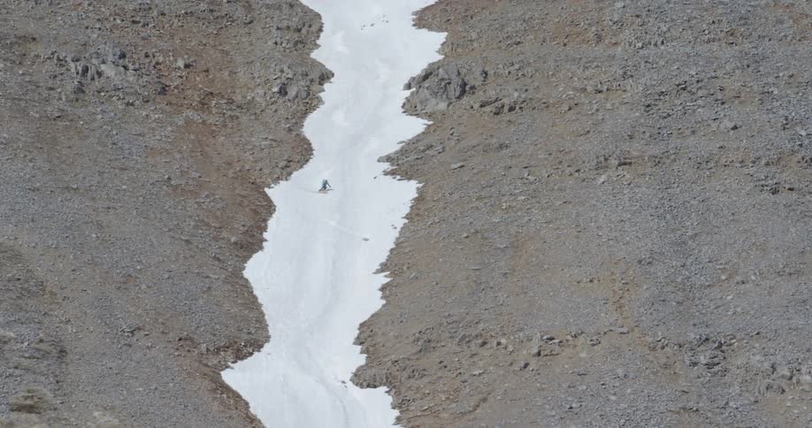 A skier skis down a strip of snow on a dry mountainside