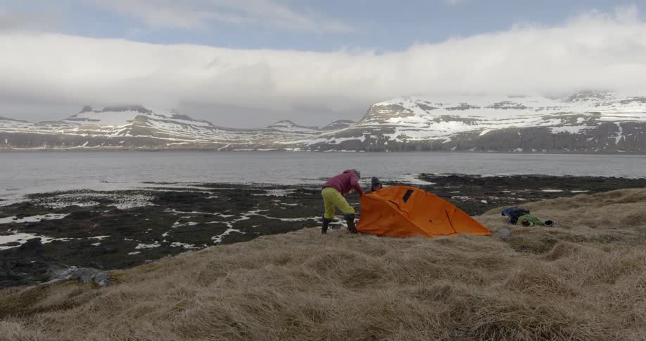 Two people putting up a tent near lake in snowy mountain wilderness