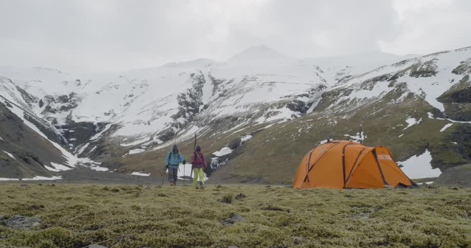 Two people hike by a tent in the mountains with skis on their back