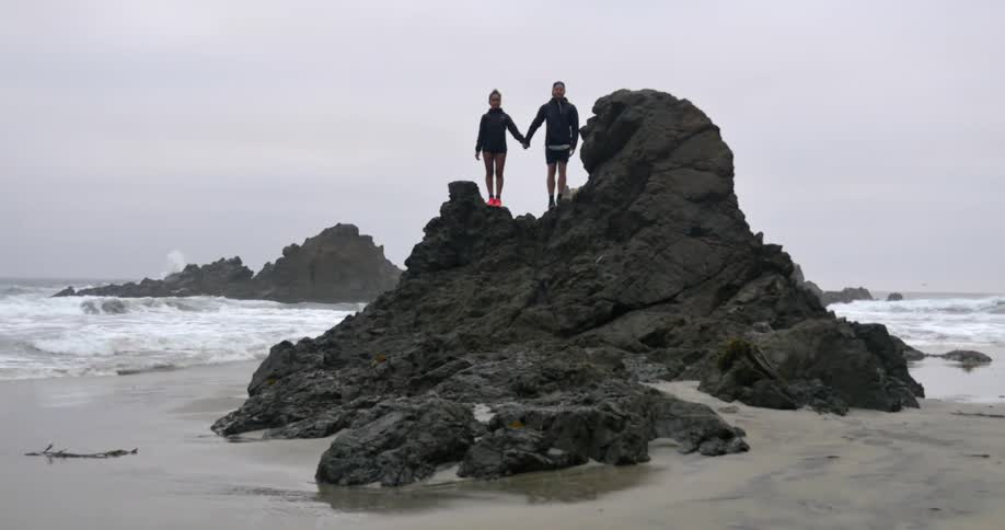 A man and woman hold hands while standing on a large rock on a beach on an overcast day