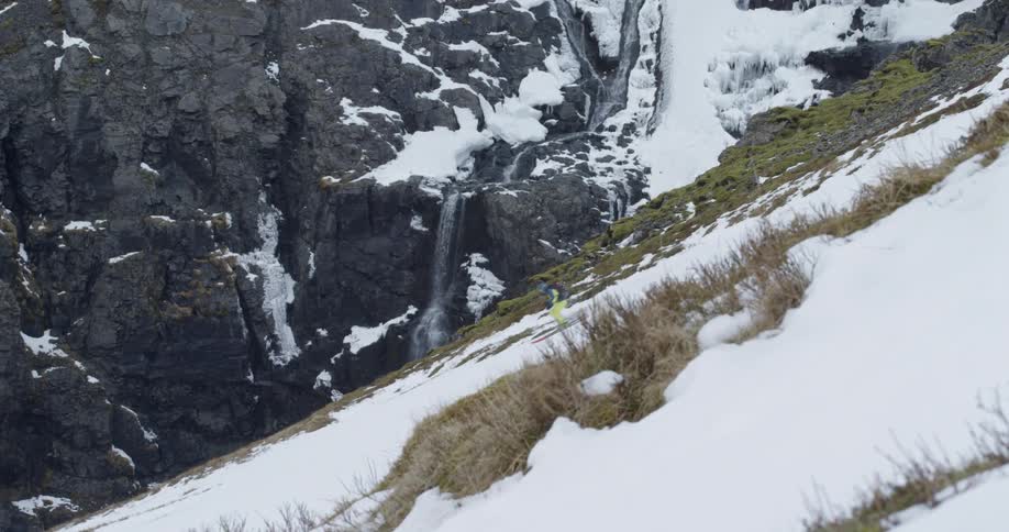 A skier crosses the frame while skiing past a waterfall