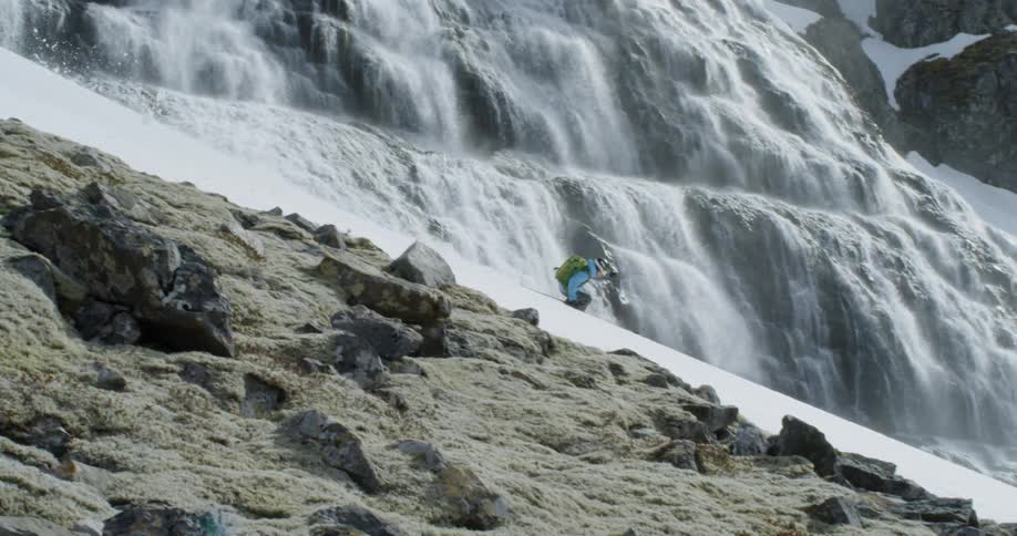 A person wearing a backpack skis down a rocky slope in front of a waterfall