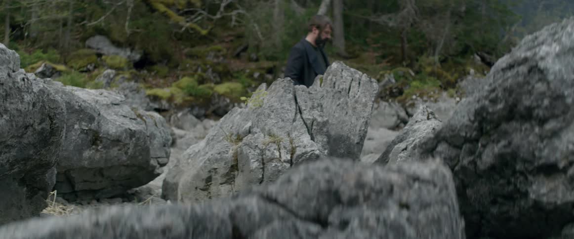A man with his hands in his coat pocket is walking by large rocks