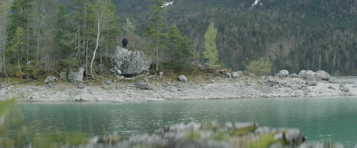 A man is standing on a large rock overlooking a lake