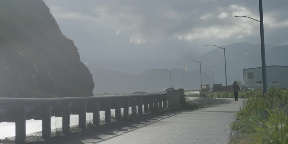 A man is walking along a sidewalk under cloudy skies