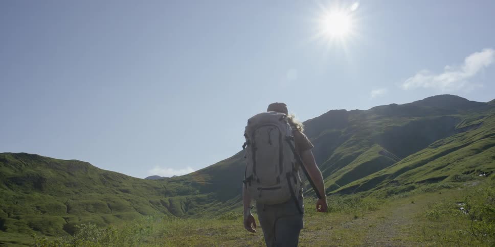 A person with a backpack is hiking down a path surrounded by green hills