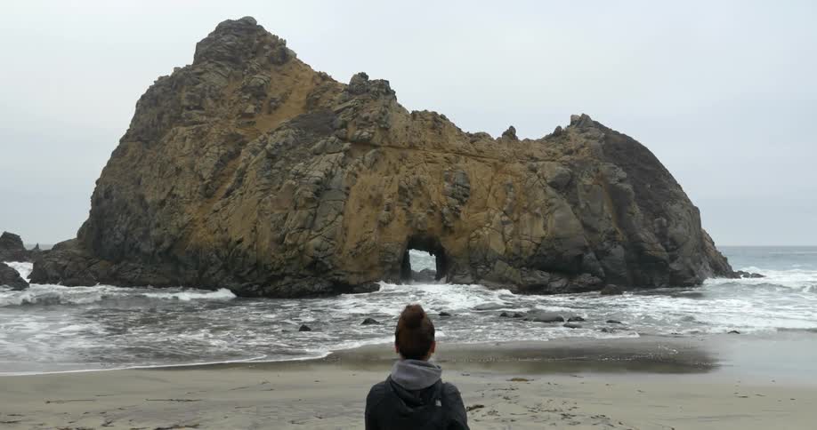 A woman on the beach looks at a rocky islet right off the coast