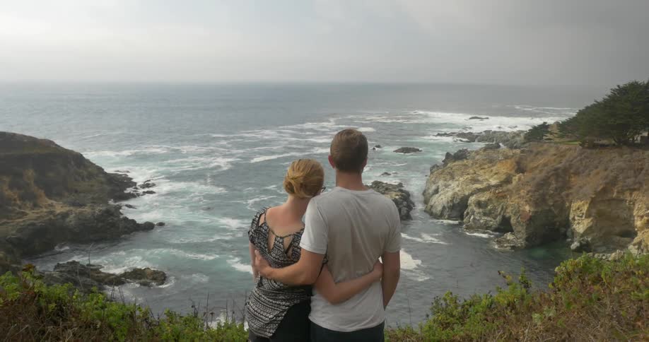 A man and woman holding each other as the lookout at rough waves under a cloudy sky