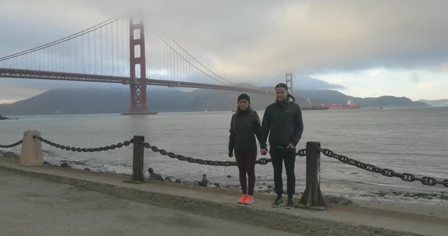 A man and woman hold hands standing next to a large river and bridge