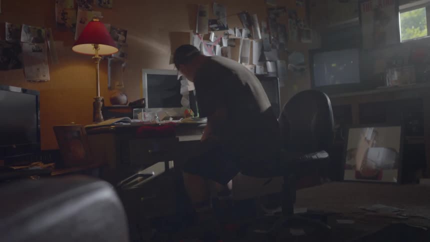A man sits down at a computer desk with papers pinned to a cork board