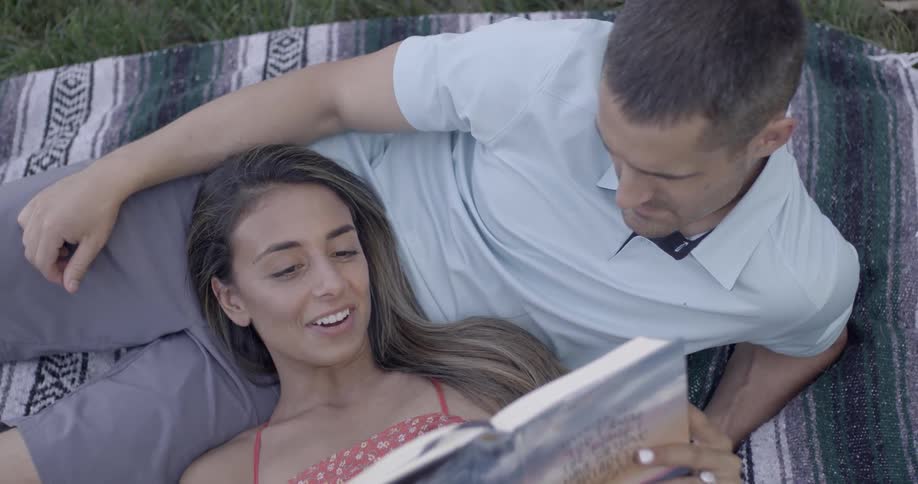 Woman reading aloud as she's resting her head on her man's lap while they lie on a blanket in the grass