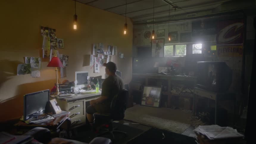 A man is sitting at a desk with a computer monitor and looks around at pictures on the wall