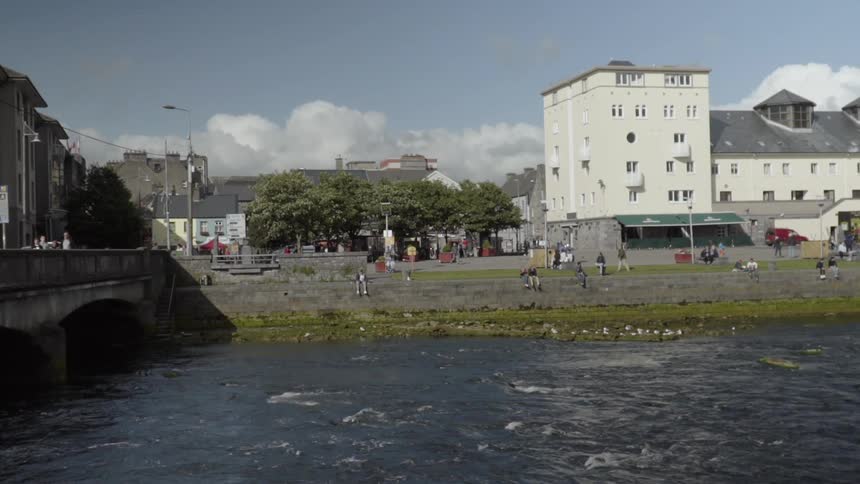 People sit on a wall watching the river flow by while people walk over the bridge