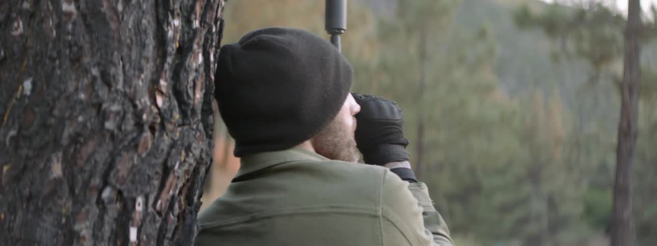 Man hiding behind a tree and then turning around to fire a rifle before retreating behind the tree again