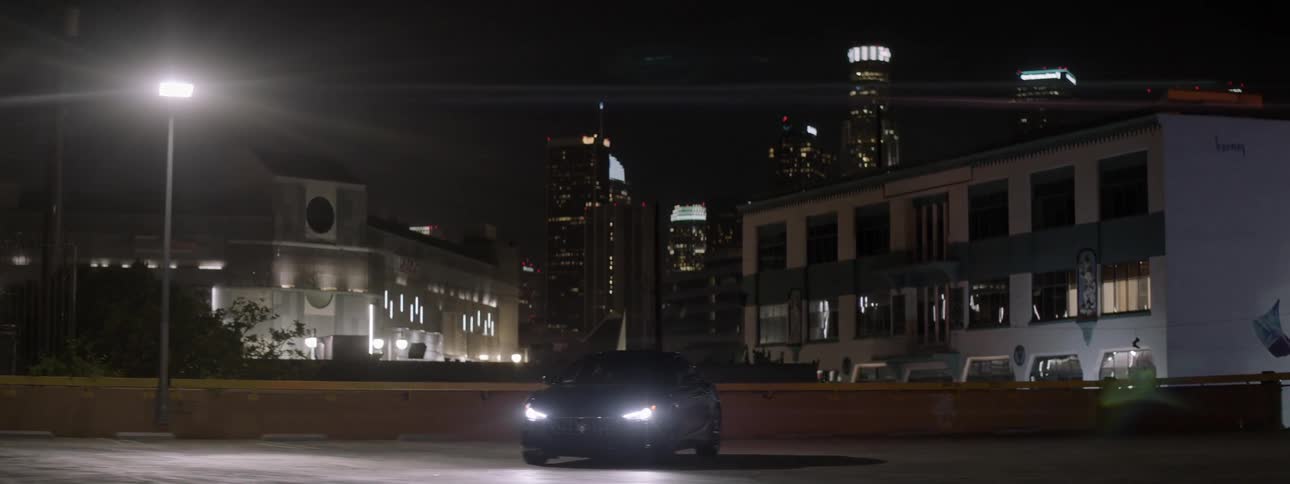 A car with its lights on is sitting on top of a parking garage with a city in the background