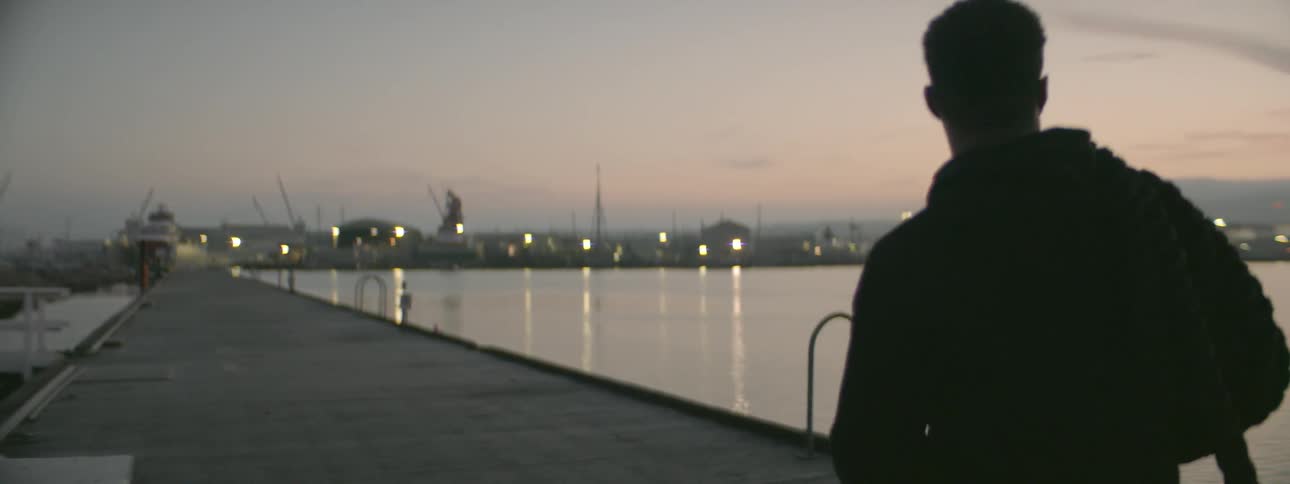 A man is carrying a heavy rope on his shoulders as he walks along a dock at dusk