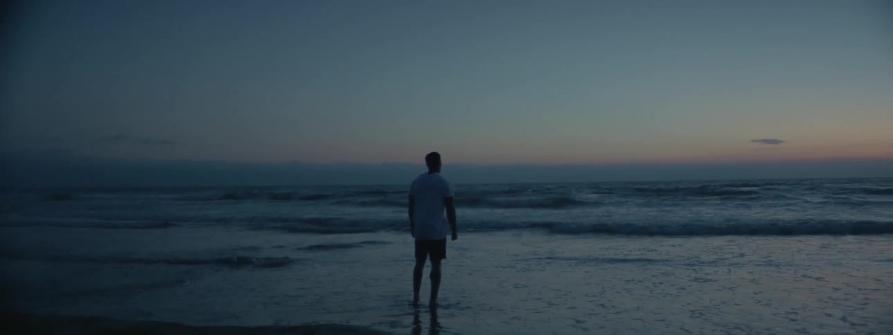 A man is standing at the ocean's edge watching the waves roll in at dusk