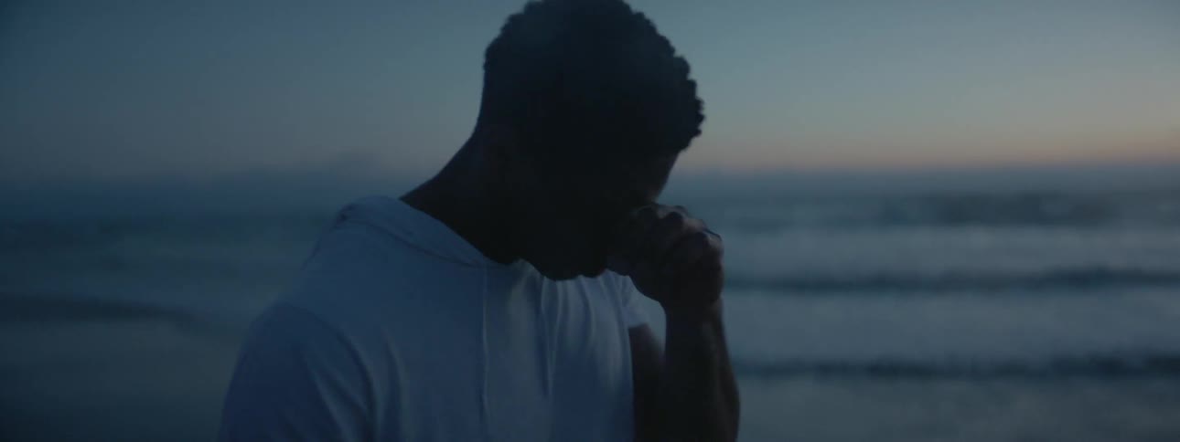 A man rubs his face while standing on the beach at sunset