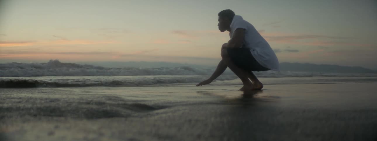 A man crouches on a beach as waves flow around him