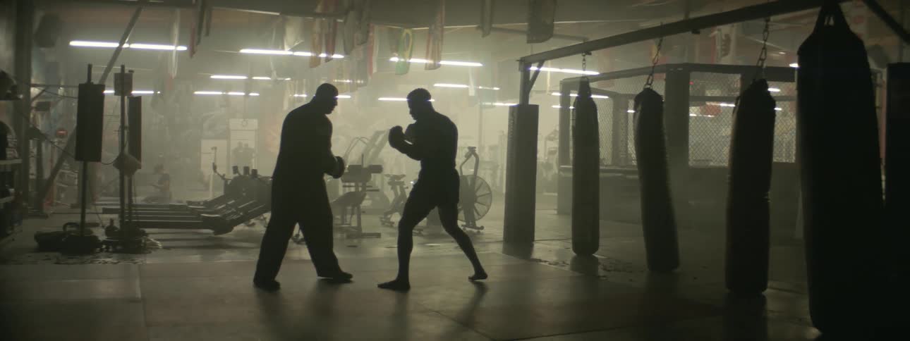 A fighter and his coach are training in a gym during the day
