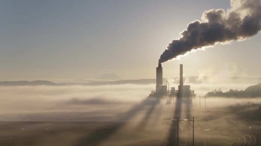 Flying along high voltage lines as the sun peaks out from beneath smoke and steam from a backlit power plant casting long shadows on the foggy ground