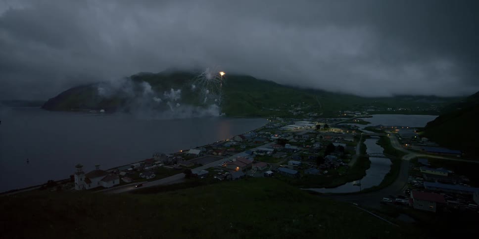 Fireworks are exploding in the sky over a coastal community on a cloudy evening