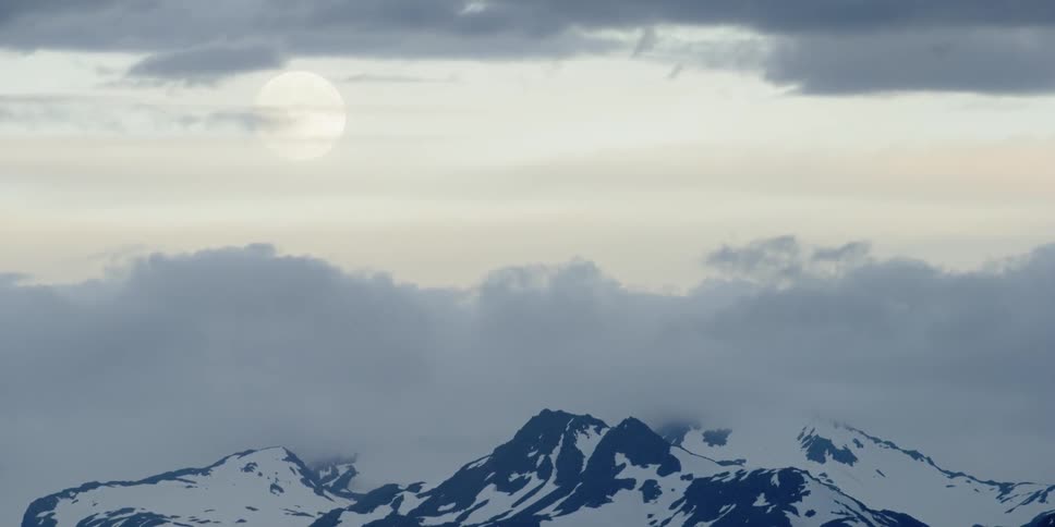 Clouds move across the moon above snow-covered mountains at dawn