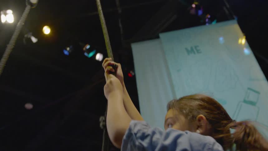 A girl tries to climb a knotted rope in an indoor arena