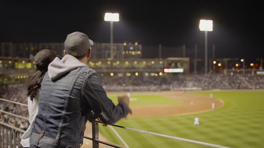 Couple leaning on a rail and tooting on their team at a night baseball game