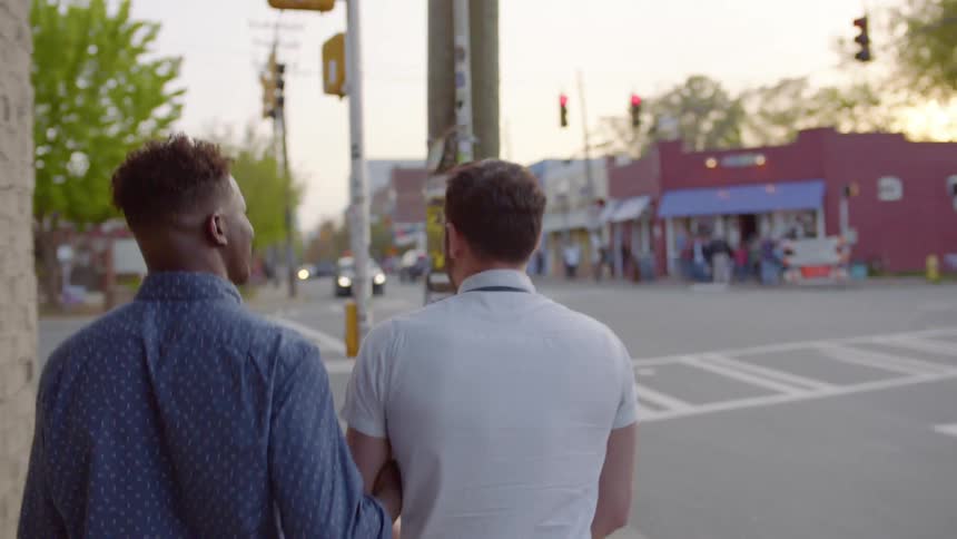 Two men walk arm and arm along a city sidewalk at dusk