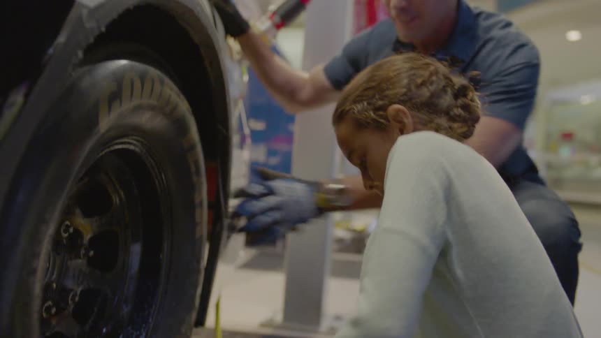A man cheers on a girl as she practices removing and resetting a tire on a race car