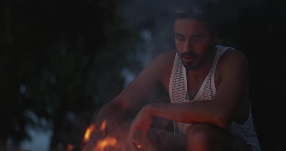 A man is holding a burning stick from a campfire at dusk