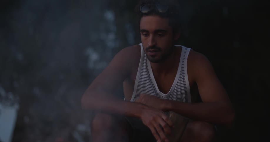 A man is sitting by a campfire at dusk preparing to add wood to it