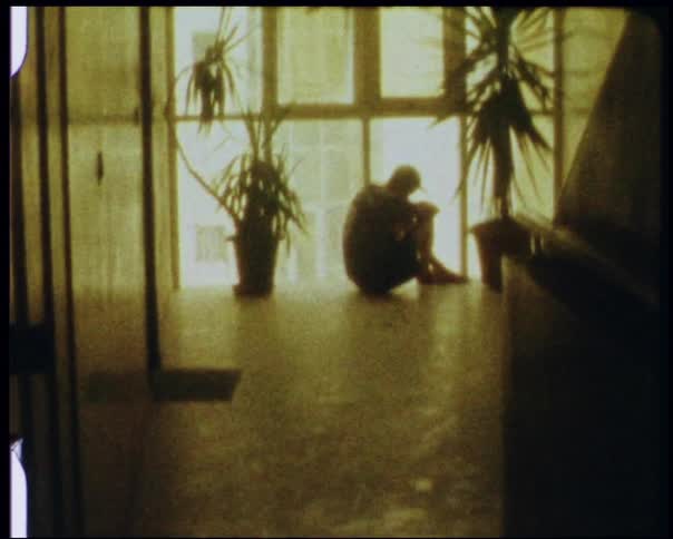 A man is sitting with his head on his knees in front of some windows during the day