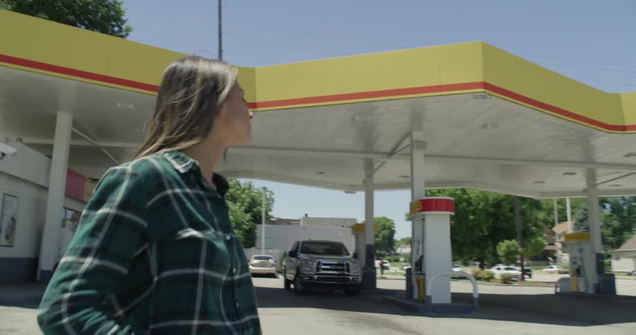 A woman is walking away from a gas station on a sunny day