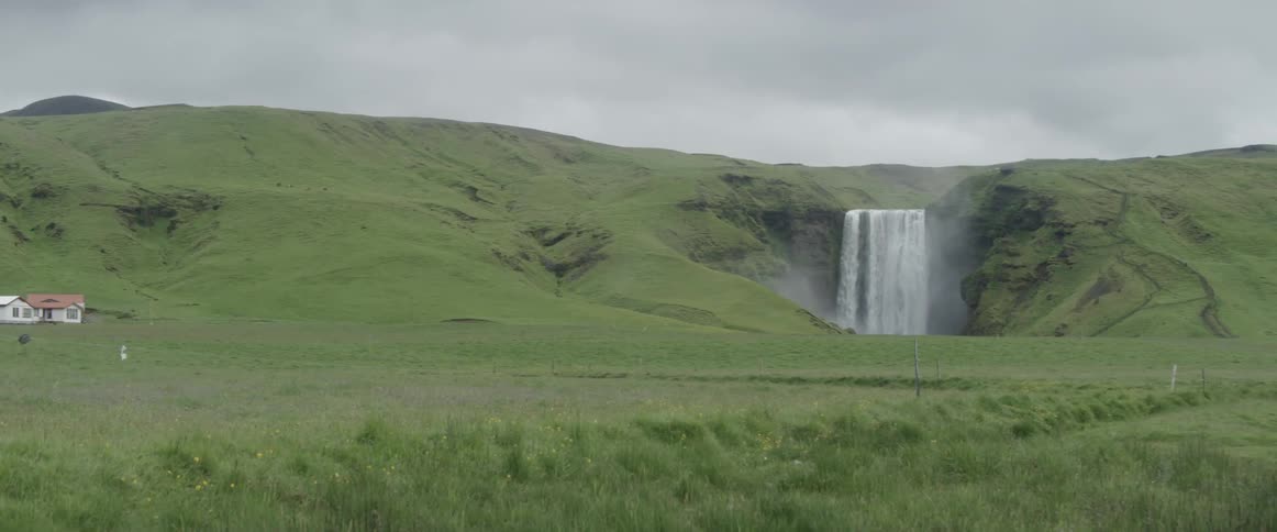 A waterfall is cascading over a cliff into a pool at the base of hills on a cloudy day