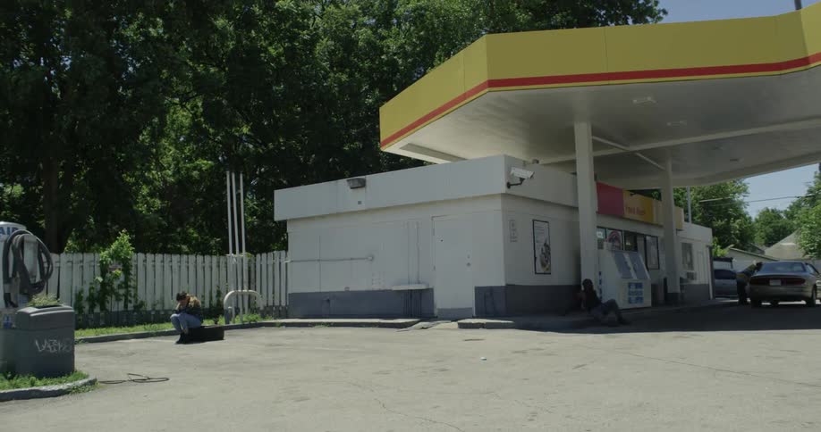 A woman is sitting on a box at the side of gas station on a sunny day