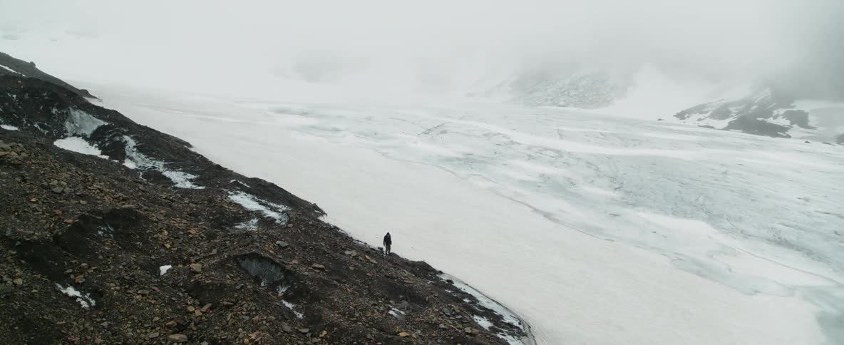A man stands at the edge of a glacier and looks at the ground on a cloudy day