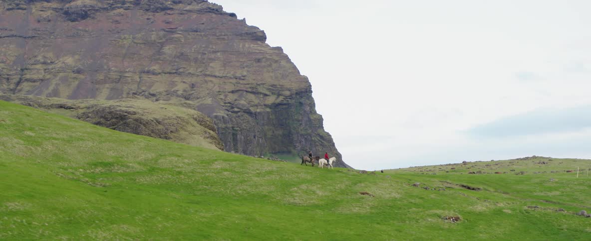 Two people are riding on horseback over a green field with a large mountain in the background