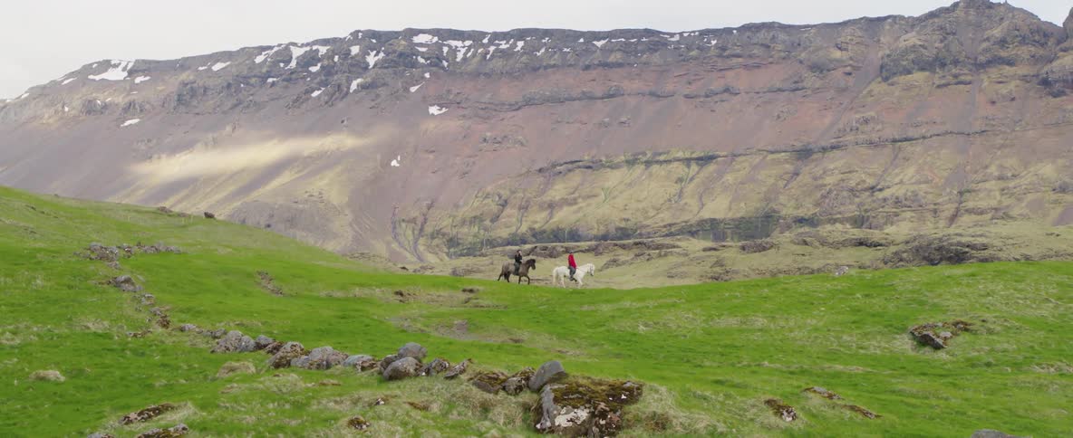 Two people ride horses near a mountain