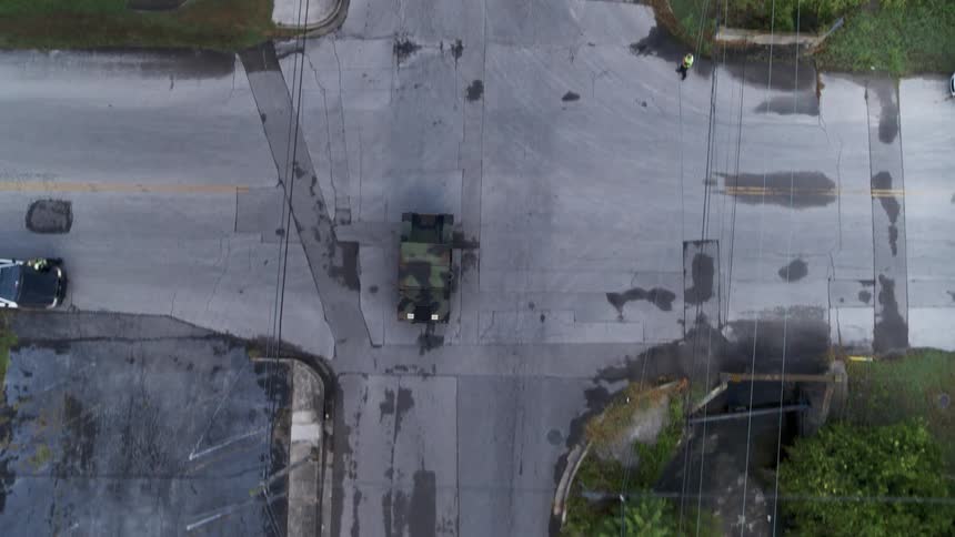 A military vehicle is driving along a rural town road during the day