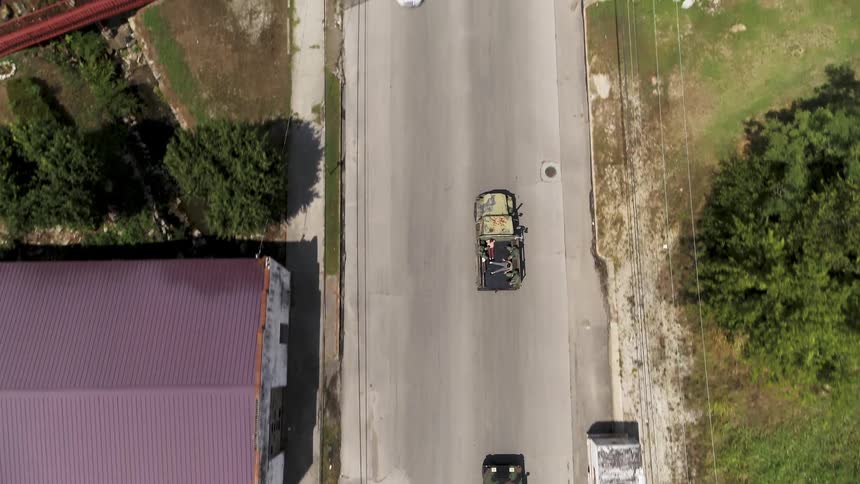 A convoy of military style vehicles is led through a town by a police car during the day